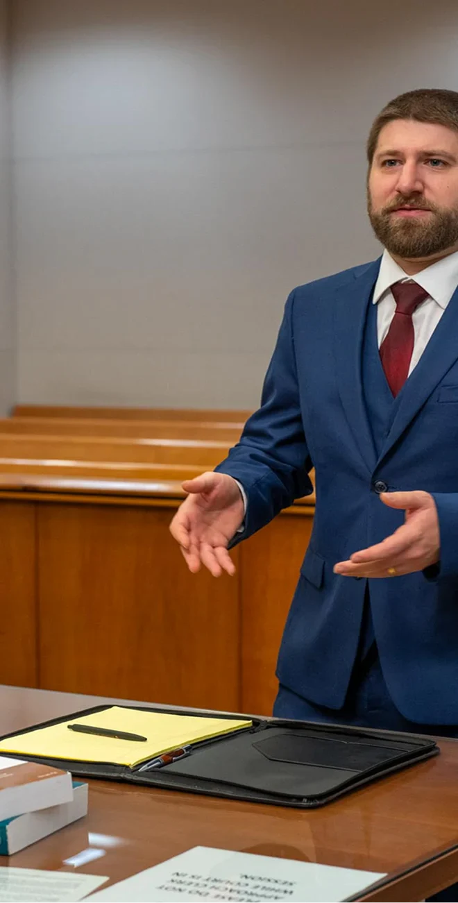 Louis Casadia in a blue suit stands at a courtroom table, gesturing, while a seated man in a gray blazer listens. Legal books and documents are visible