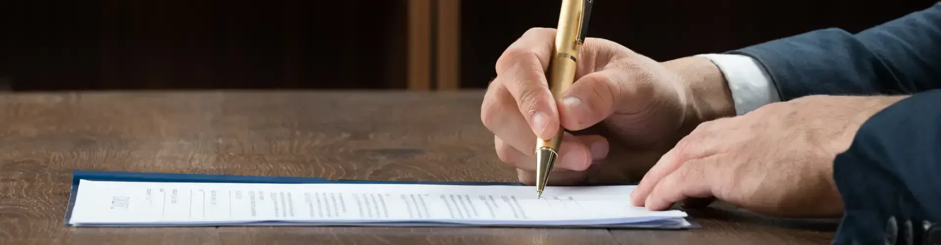 A person's hand, holding a gold pen, signs a document on a wooden table, focusing on a sheet of paper with text.